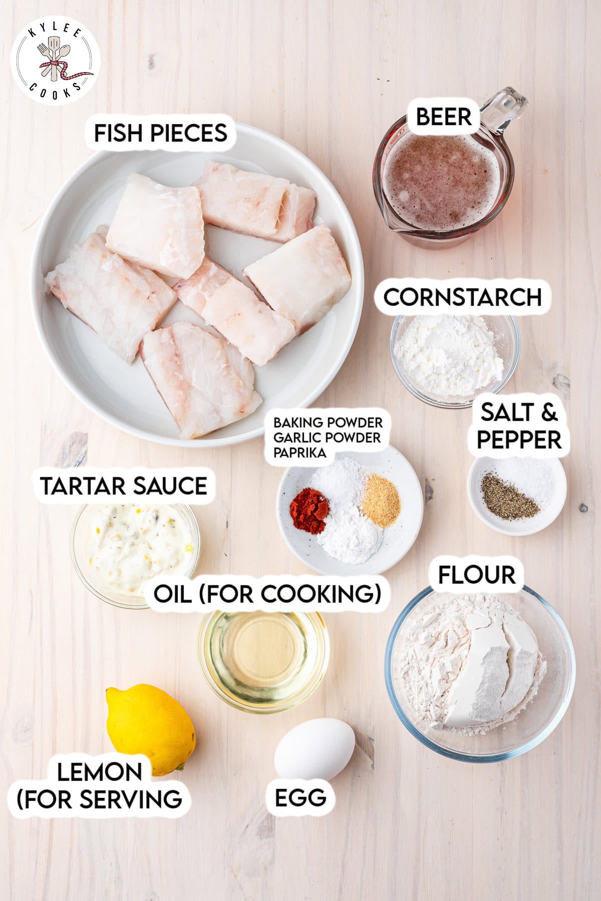 A flat lay of ingredients for fish and chips: fish pieces, beer, cornstarch, salt, pepper, flour, egg, oil, tartar sauce, a lemon, and spices on a light surface.