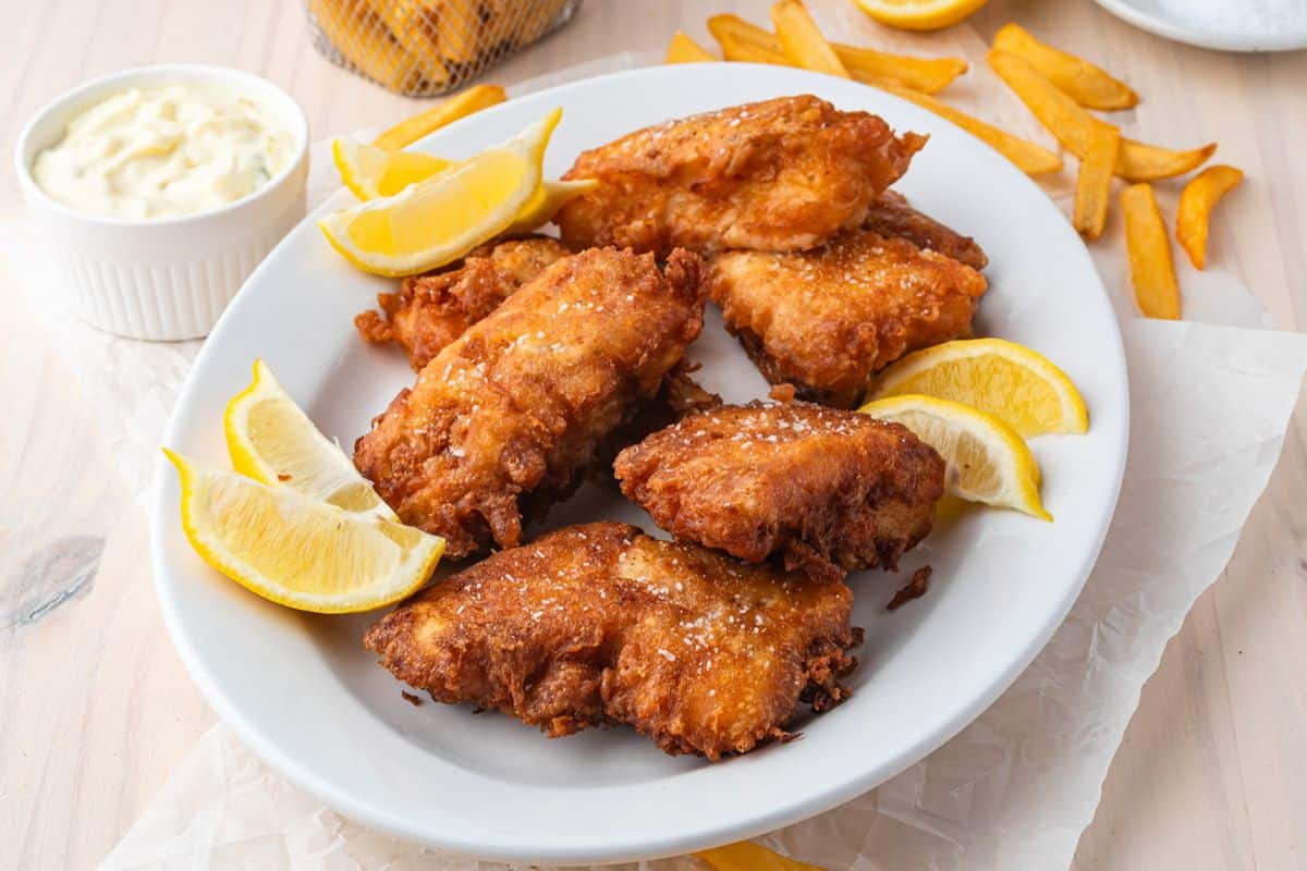 A white plate with fried fish fillets, lemon wedges, and a side of French fries. A small bowl of tartar sauce is visible in the background.