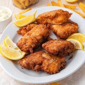 A white plate with several pieces of golden-brown fried fish, garnished with lemon wedges, with fries and tartar sauce in the background.
