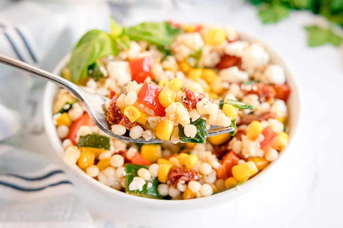A close-up of a spoonful of Summer Couscous Salad with corn, spinach, tomatoes, and cheese, held above a bowl filled with more vibrant salad.