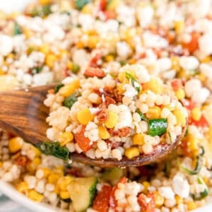 A wooden spoon holds a serving of Summer Couscous Salad with corn, zucchini, red peppers, and crumbled cheese.