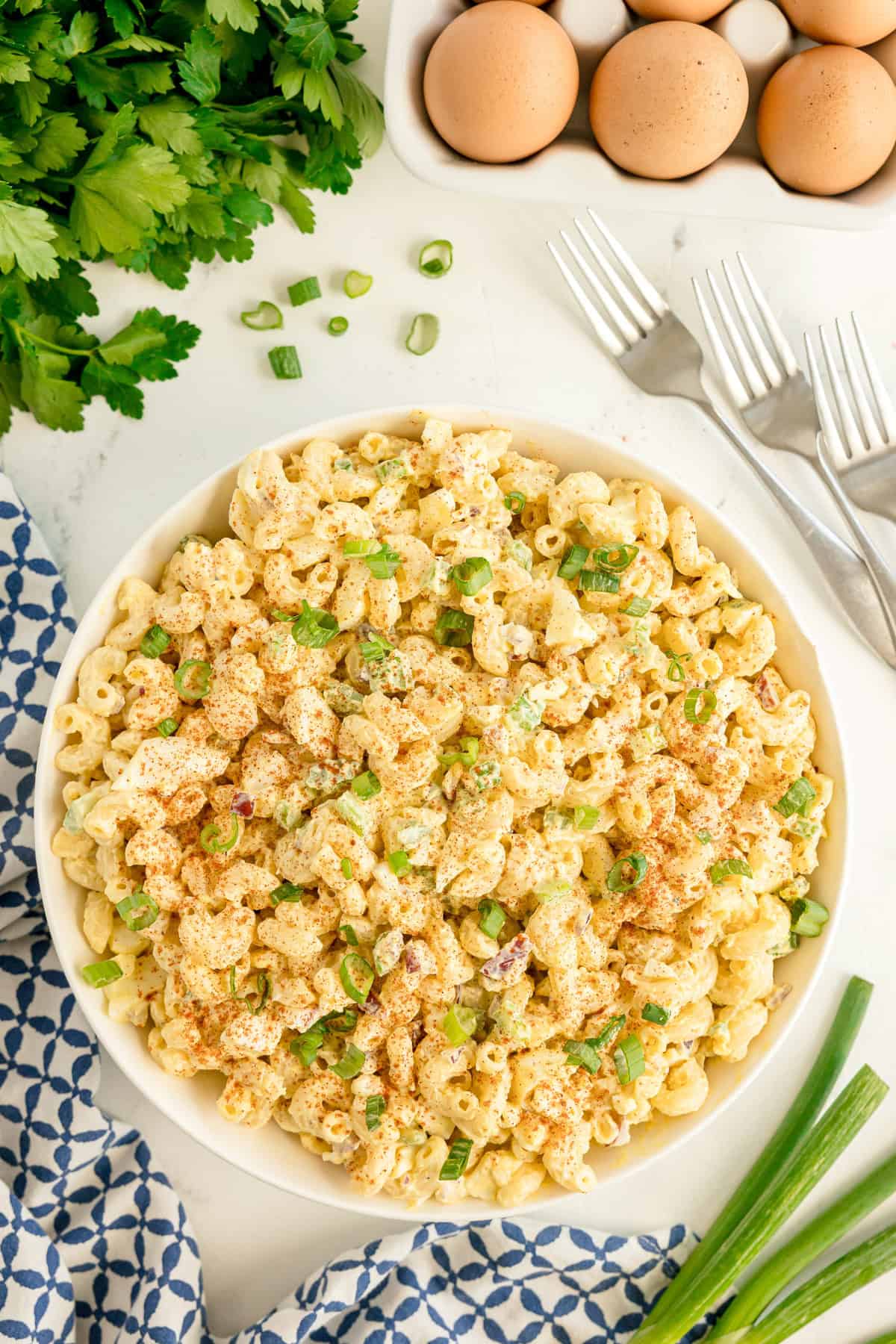 A bowl of Deviled Egg Pasta Salad garnished with chopped green onions and paprika, surrounded by parsley, eggs, and forks on a white table. This easy recipe is perfect as a make ahead recipe for gatherings or meal prep.