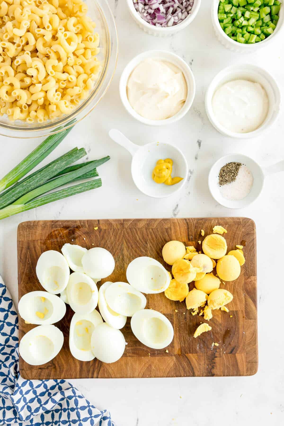 Chopped hard-boiled eggs on a wooden board, surrounded by bowls of pasta, mayonnaise, sour cream, chopped vegetables, mustard, salt, and pepper on a white countertop.