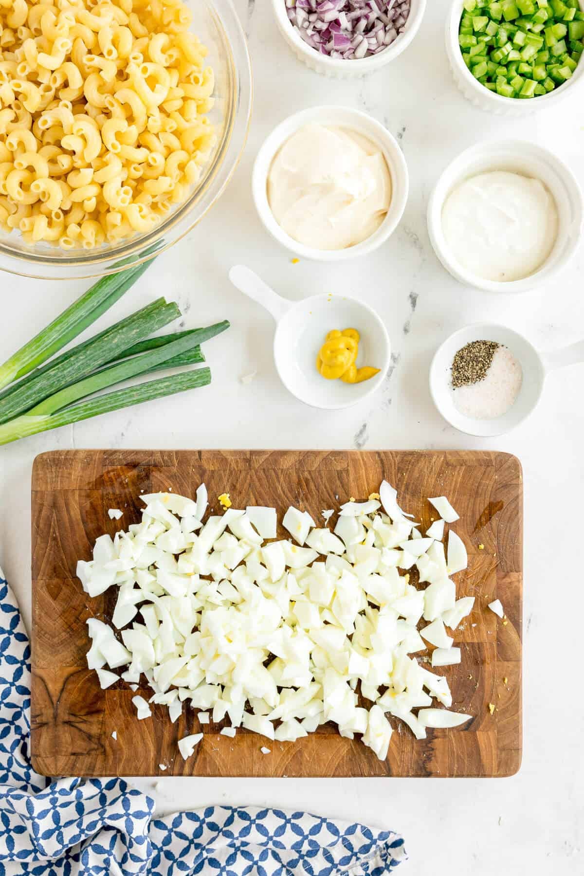 Chopped hard-boiled eggs on a wooden cutting board surrounded by uncooked macaroni, green onions, and bowls of mayonnaise, mustard, diced vegetables, salt, and pepper.