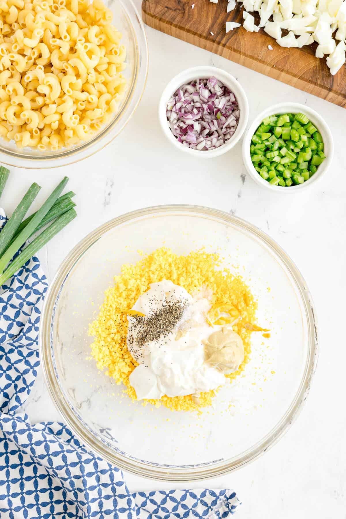 A glass bowl with egg yolks, mayonnaise, mustard, salt, and pepper, surrounded by bowls of macaroni, diced onion, green pepper, and chopped egg whites on a white countertop.