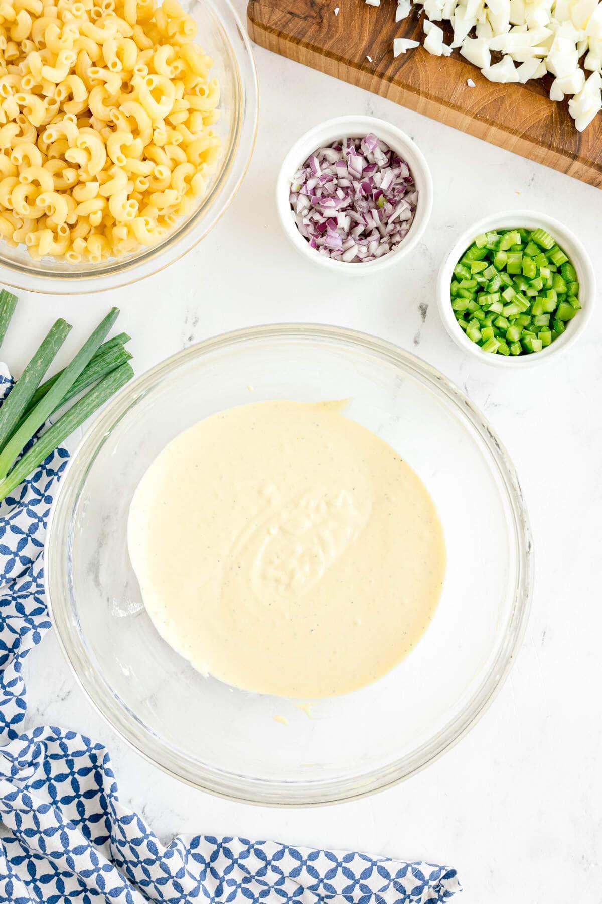 A glass bowl of creamy dressing sits beside bowls of cooked macaroni, chopped red onion, chopped green pepper, and a cutting board with chopped hard-boiled eggs.