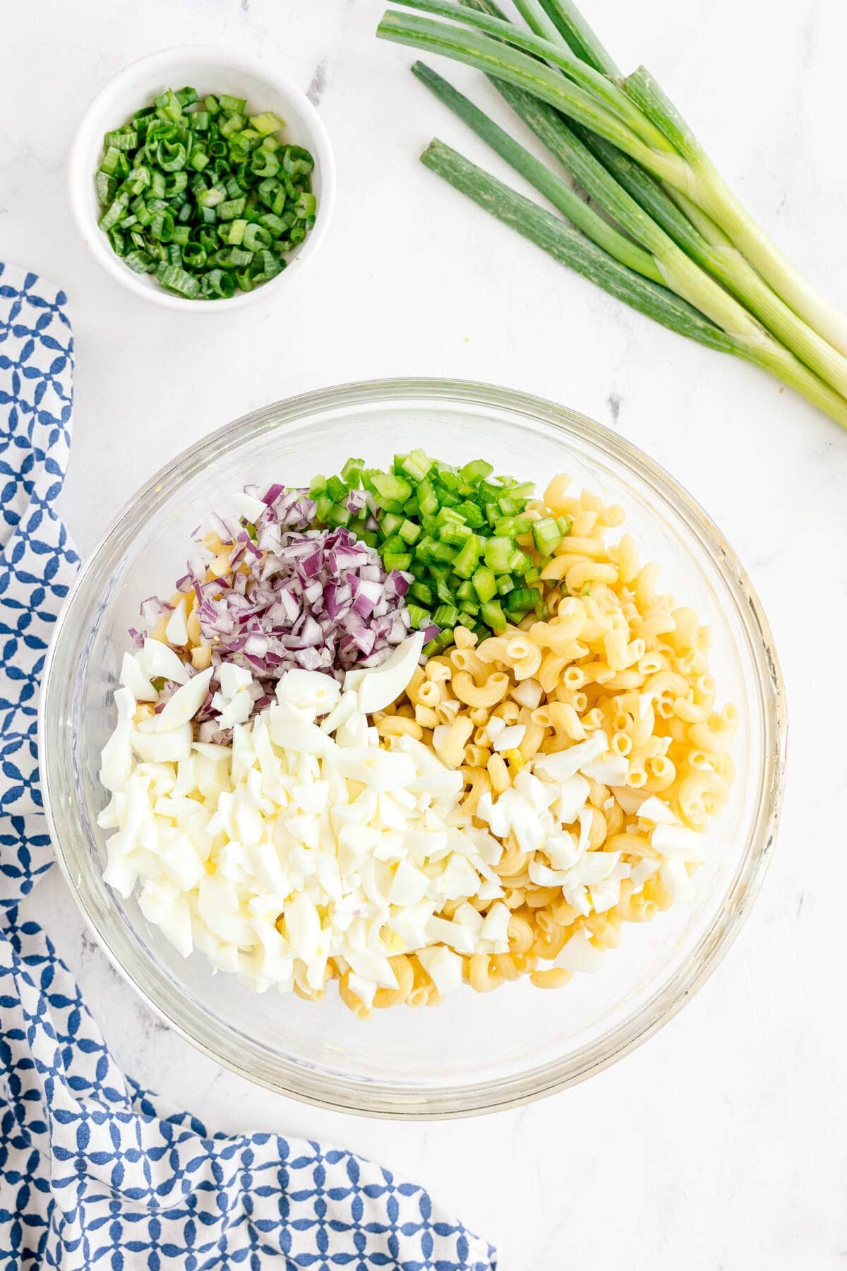 A glass bowl with cooked macaroni, chopped hard-boiled eggs, red onion, and green bell pepper, next to green onions and a small bowl of chopped green onions on a white surface.