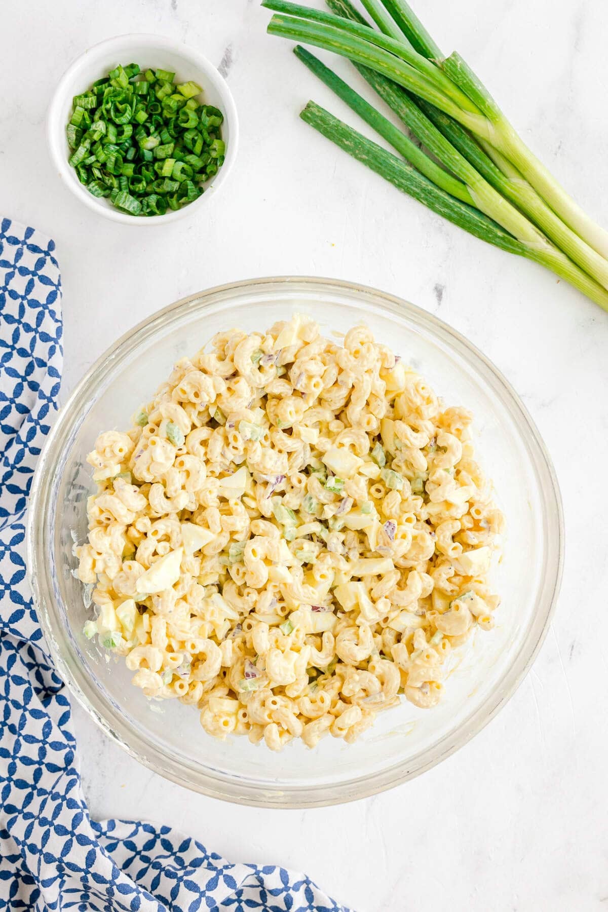 A glass bowl of macaroni salad on a white surface, with a bunch of green onions, a small bowl of chopped green onions, and a blue-patterned towel nearby.