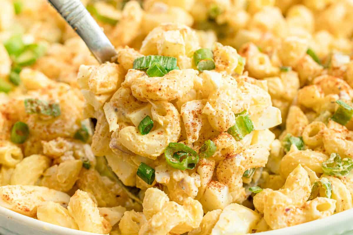 A close-up of a serving spoon scooping macaroni salad with chopped green onions and a light sprinkle of paprika.