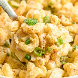 Close-up of macaroni salad with chopped green onions, egg pieces, and sprinkled paprika, served with a spoon.