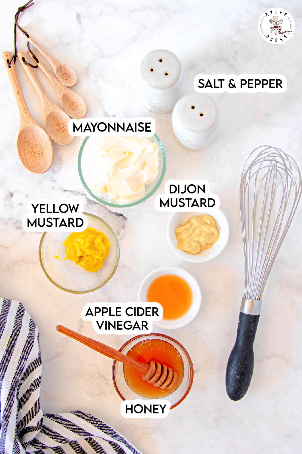 Overhead view of ingredients for a Honey Mustard Sauce: mayonnaise, yellow mustard, Dijon mustard, apple cider vinegar, honey, salt, pepper, wooden spoons, and a whisk on a white surface.