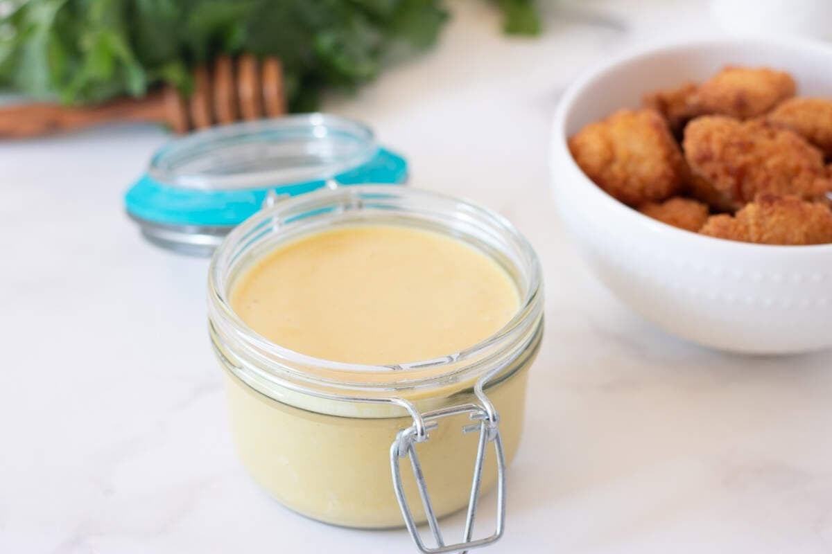 A glass jar filled with honey mustard sauce sits on a white surface, ready to be used as a dipping sauce for the bowl of breaded chicken nuggets and leafy greens in the background.