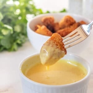A fork holds a chicken nugget dipped in honey mustard sauce, with a bowl of more nuggets and leafy greens in the background.