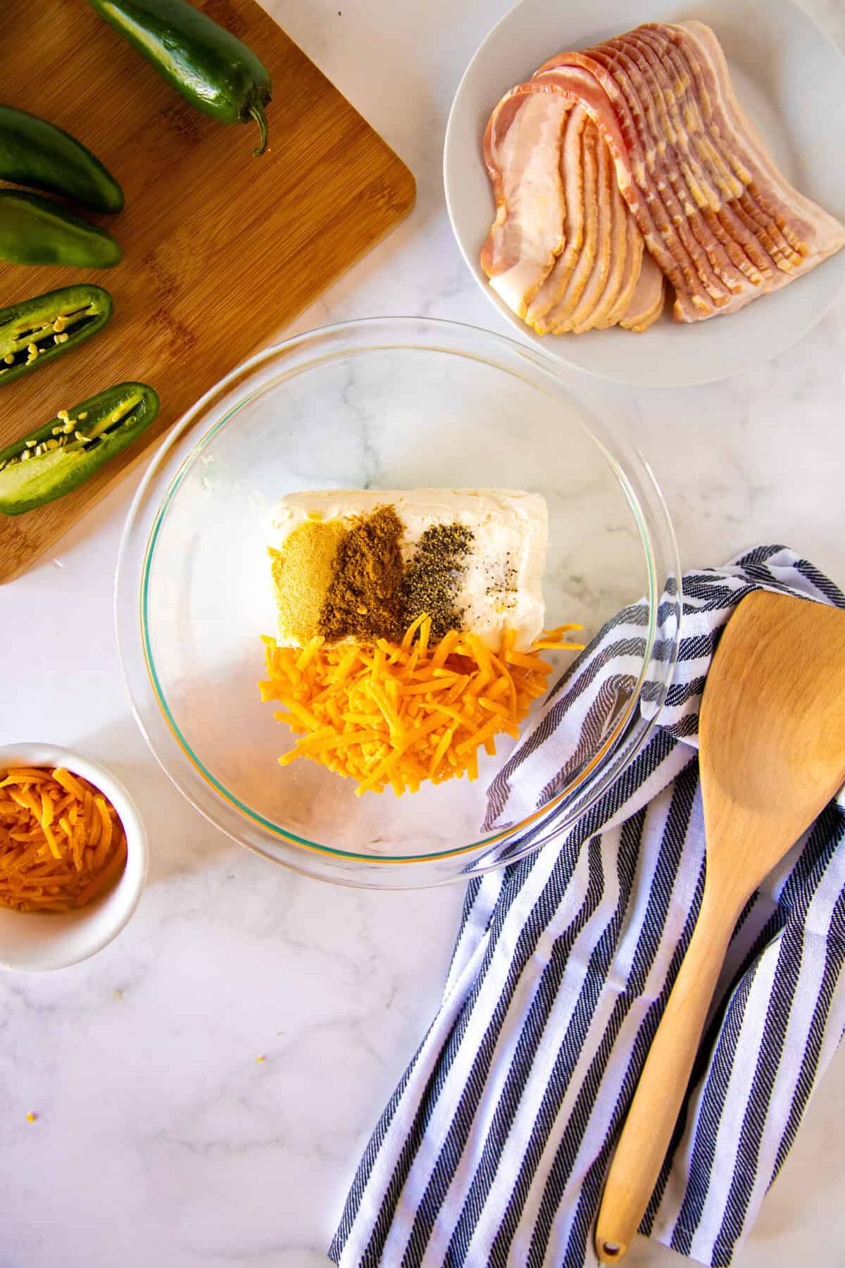 A glass bowl with shredded cheese, spices, and cream cheese, next to a striped towel, wooden spoon, jalapeños on a cutting board, bacon slices, and a small bowl of cheese.