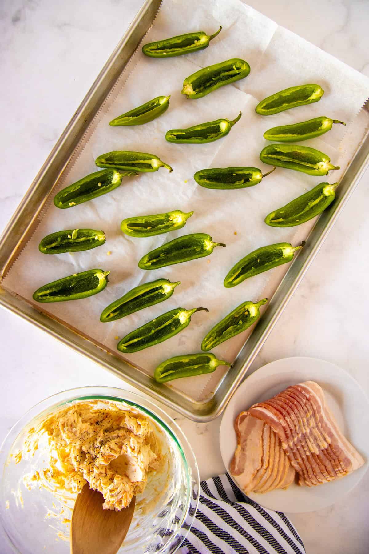 Halved jalapeños on a parchment-lined baking sheet, a bowl of cheese mixture with a wooden spoon, and raw bacon slices on a plate.