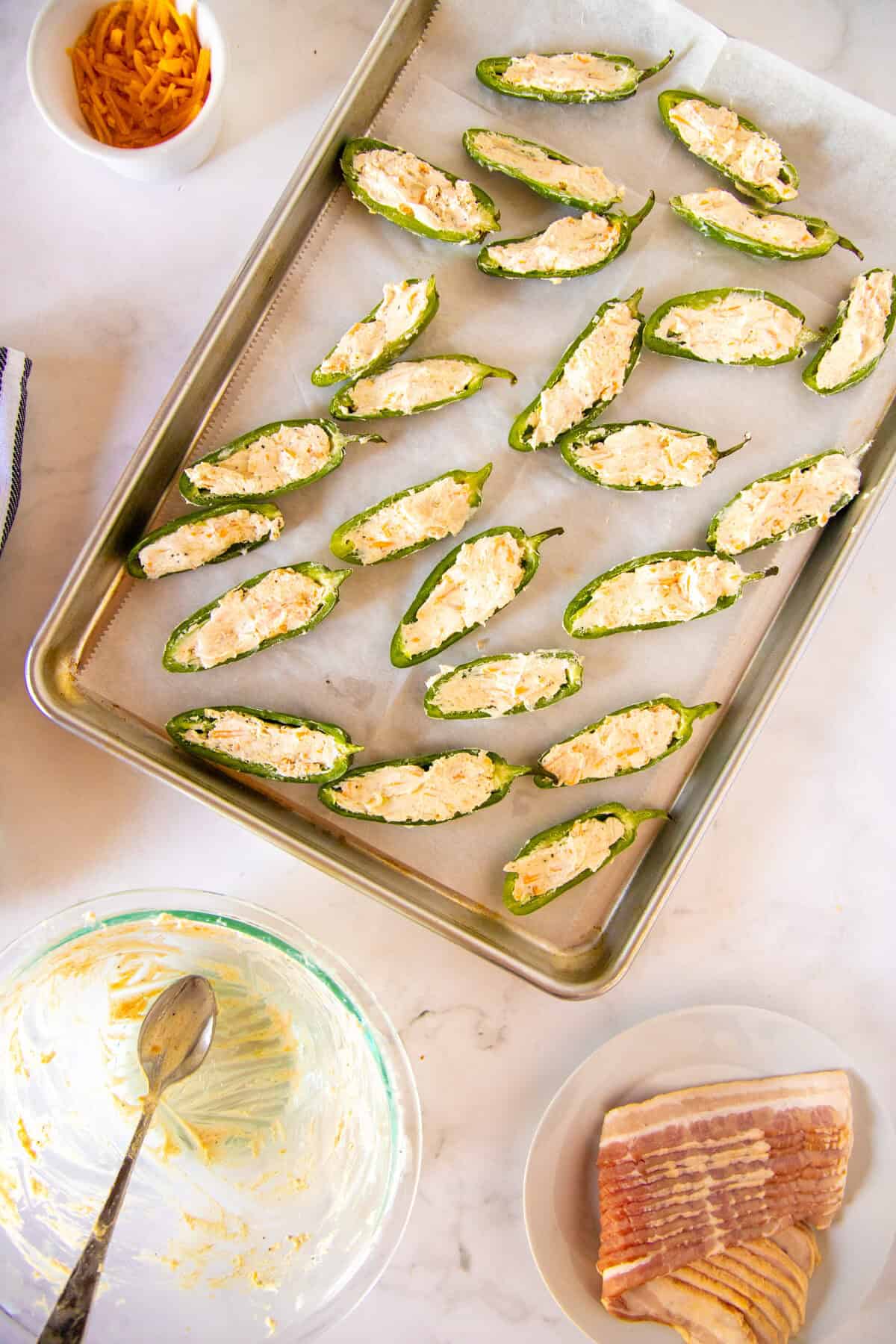 A baking sheet with halved jalapeños stuffed with cheese mixture, surrounded by a bowl of shredded cheese, a bowl of cream cheese mixture, and a plate of uncooked bacon.