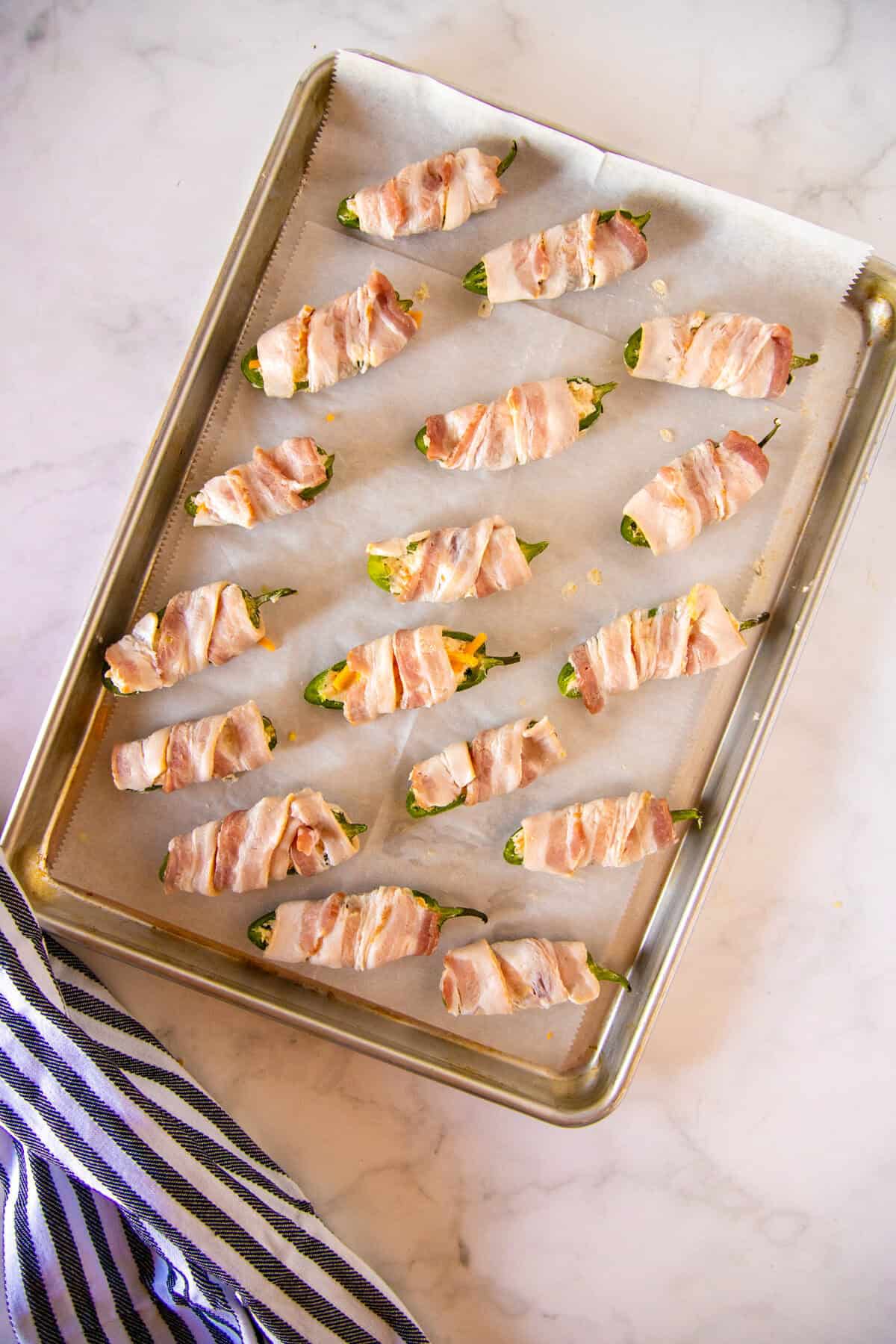 A baking sheet lined with parchment paper holds several bacon-wrapped jalapeño poppers, arranged in rows, on a marble countertop with a striped towel nearby.
