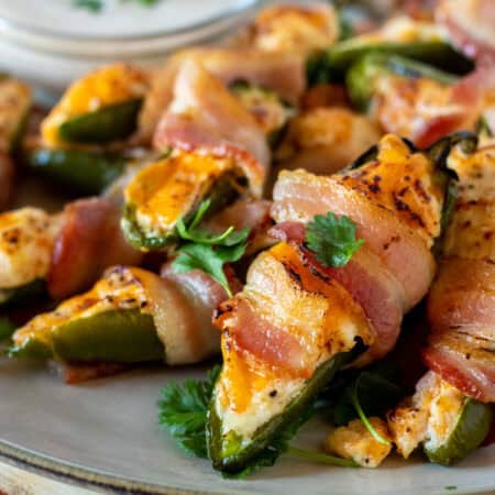 Plate of bacon-wrapped jalapeño poppers with melted cheese, garnished with herbs, and a small bowl of dipping sauce in the background.
