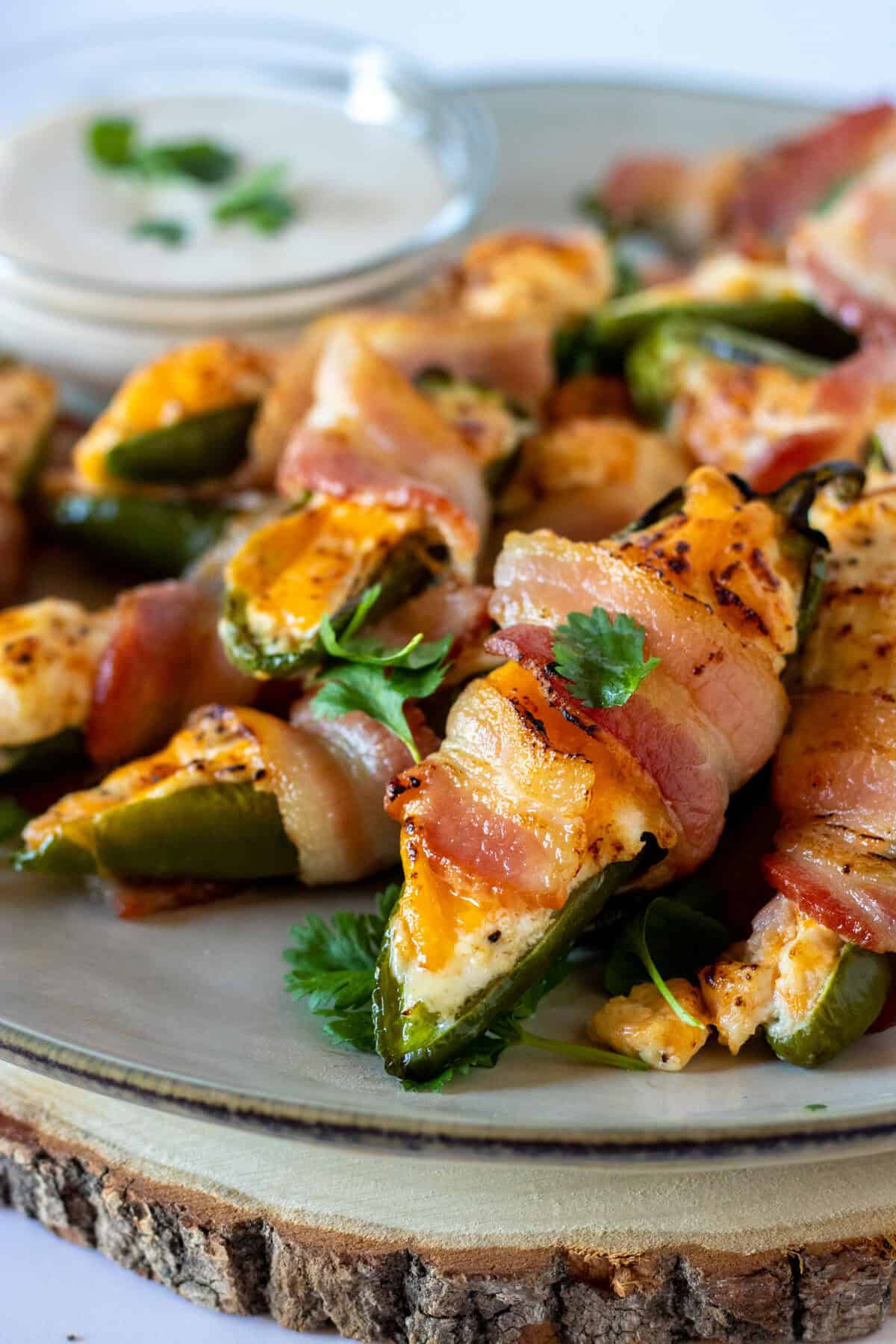 Plate of bacon-wrapped jalapeño poppers with melted cheese, garnished with herbs, and a small bowl of dipping sauce in the background.