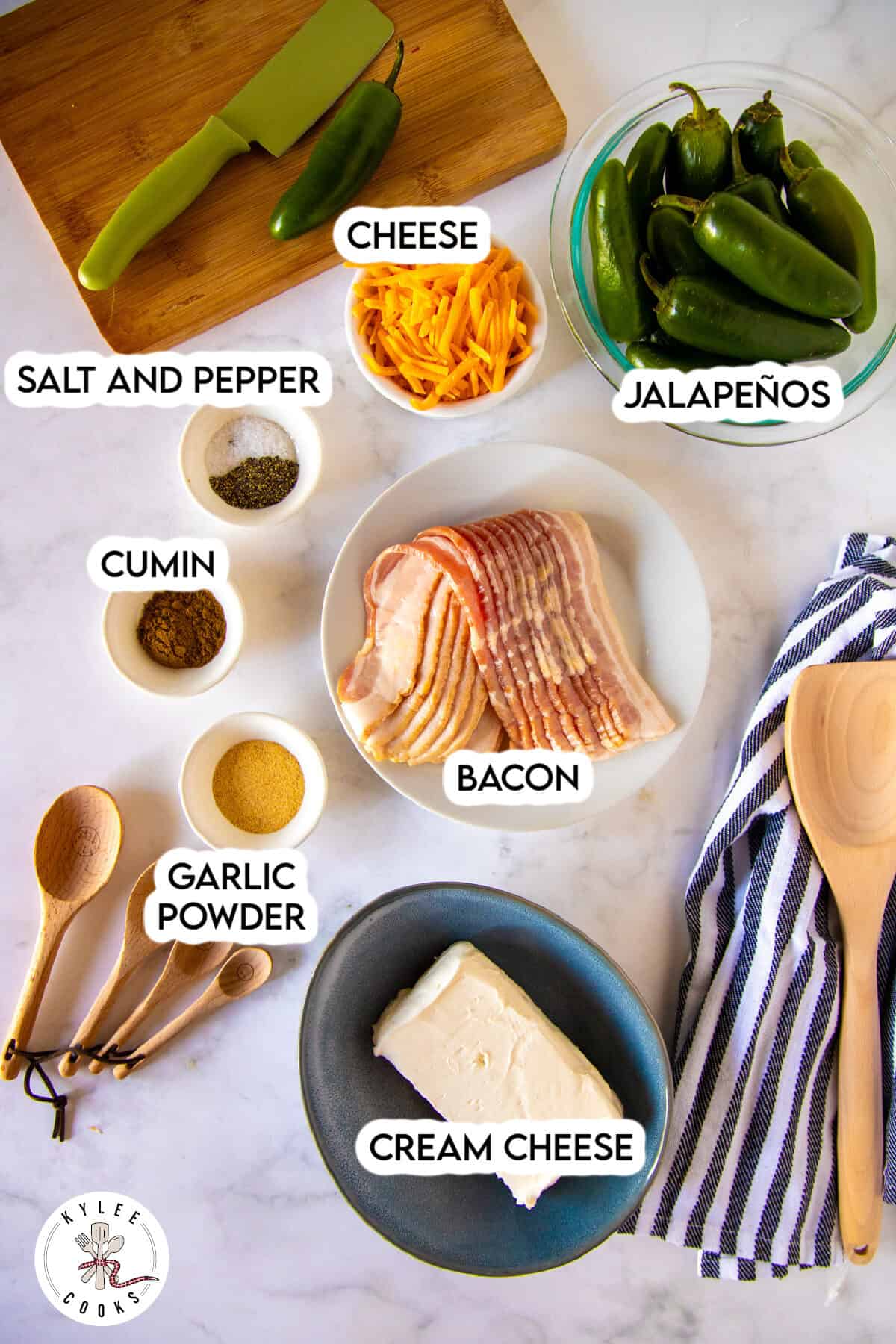 A variety of ingredients on a countertop, including bacon, shredded cheese, whole jalapeños, cream cheese, spices, and wooden utensils, all labeled.