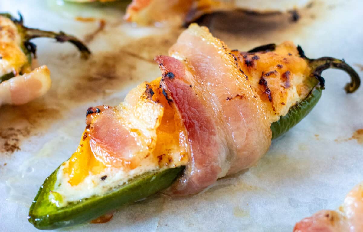 A jalapeño pepper stuffed with cheese, wrapped in bacon, and baked on a parchment-lined surface.