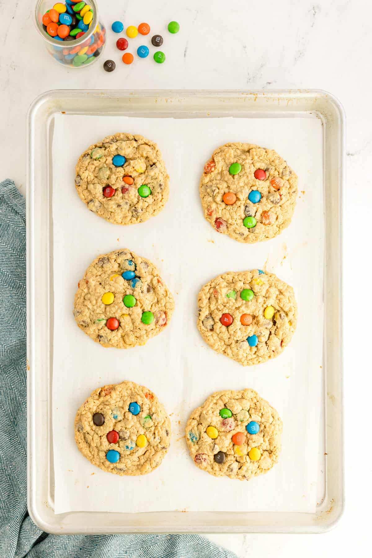 Six large oatmeal cookies topped with colorful candy-coated chocolate pieces are arranged on a parchment-lined baking sheet, with a jar of candies and a kitchen towel nearby.