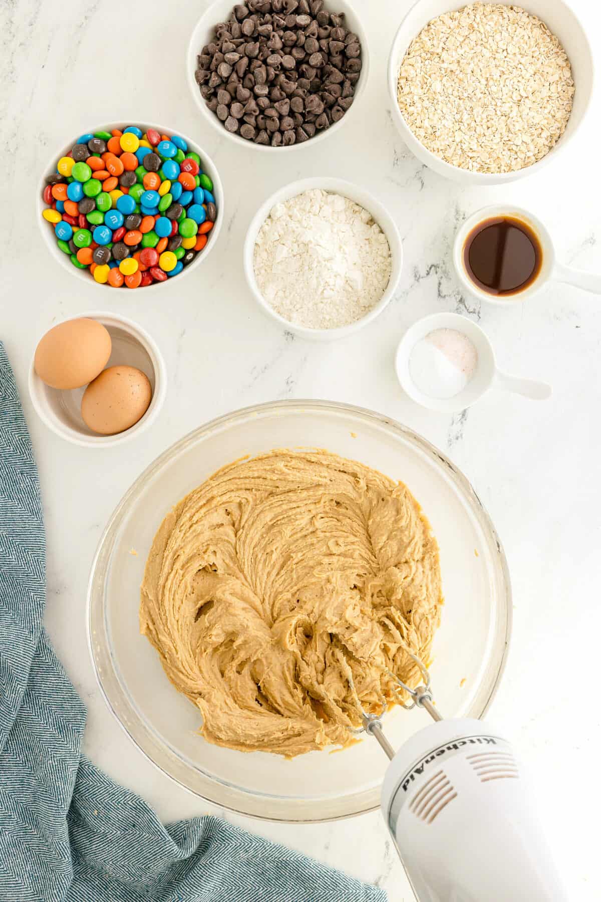 A mixing bowl with creamed dough and a hand mixer surrounded by bowls of oats, chocolate chips, M&Ms, flour, vanilla, salt, and two eggs on a white surface.