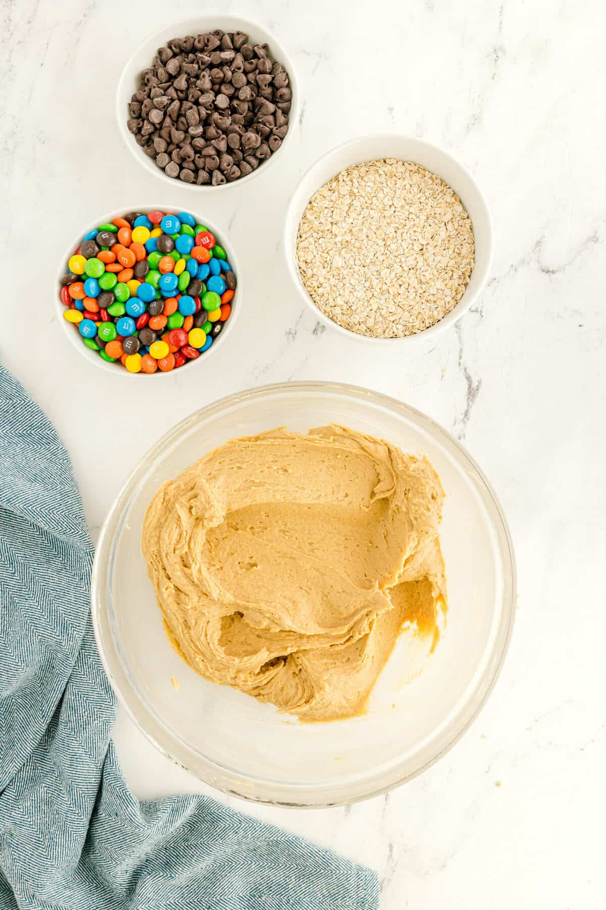 A glass bowl with cookie dough sits on a marble surface next to bowls of chocolate chips, oats, and colorful candy-coated chocolates. A blue cloth is partially visible on the left.