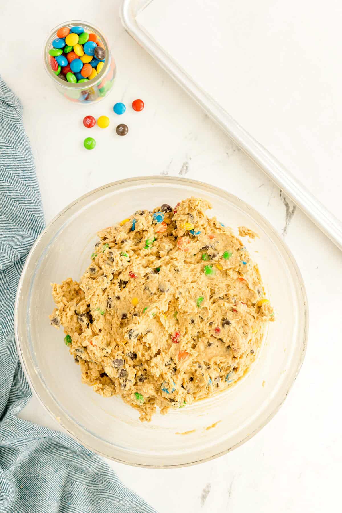 A glass bowl filled with cookie dough mixed with colorful candy-coated chocolates sits on a white countertop next to scattered candies and a blue cloth.