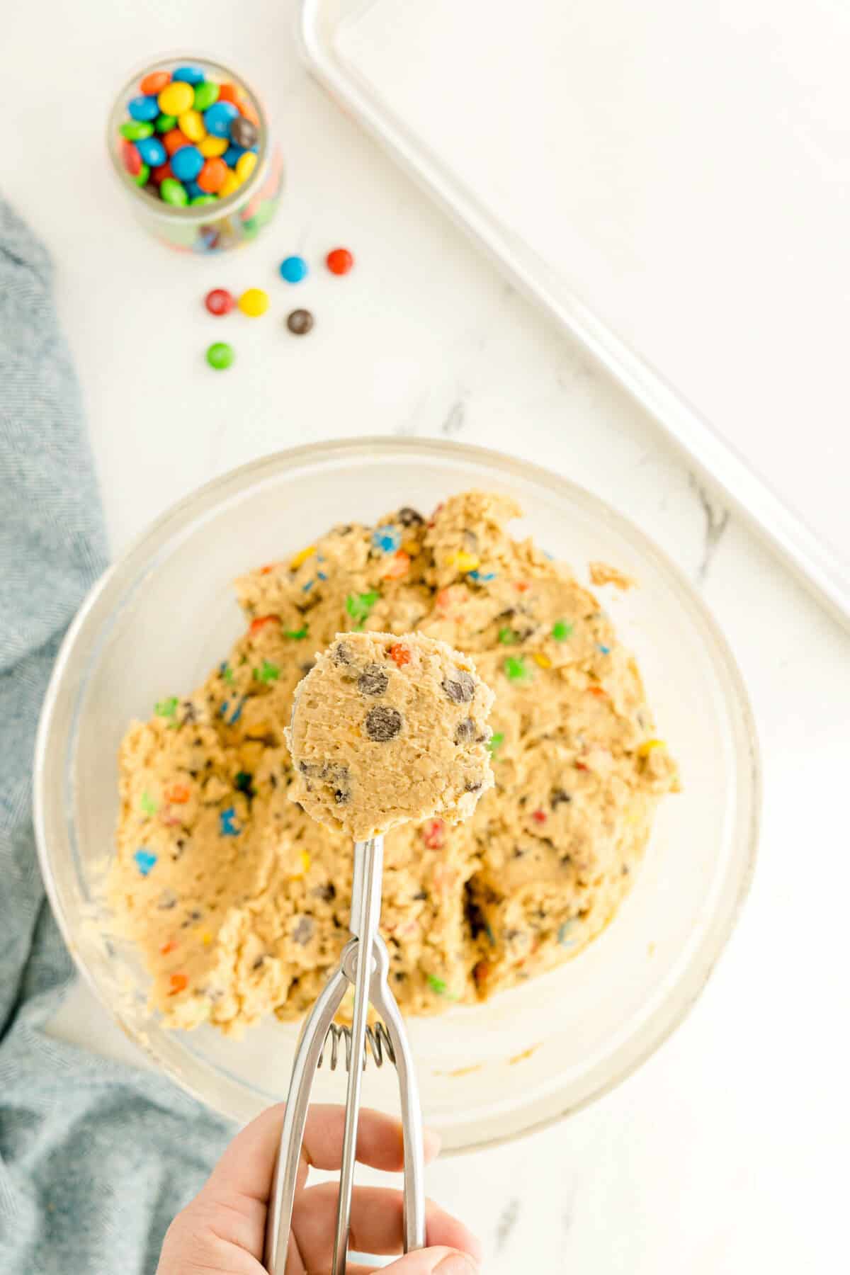 A hand holds a cookie scoop with dough over a bowl of cookie dough mixed with colorful candies; a baking tray and candies are nearby.