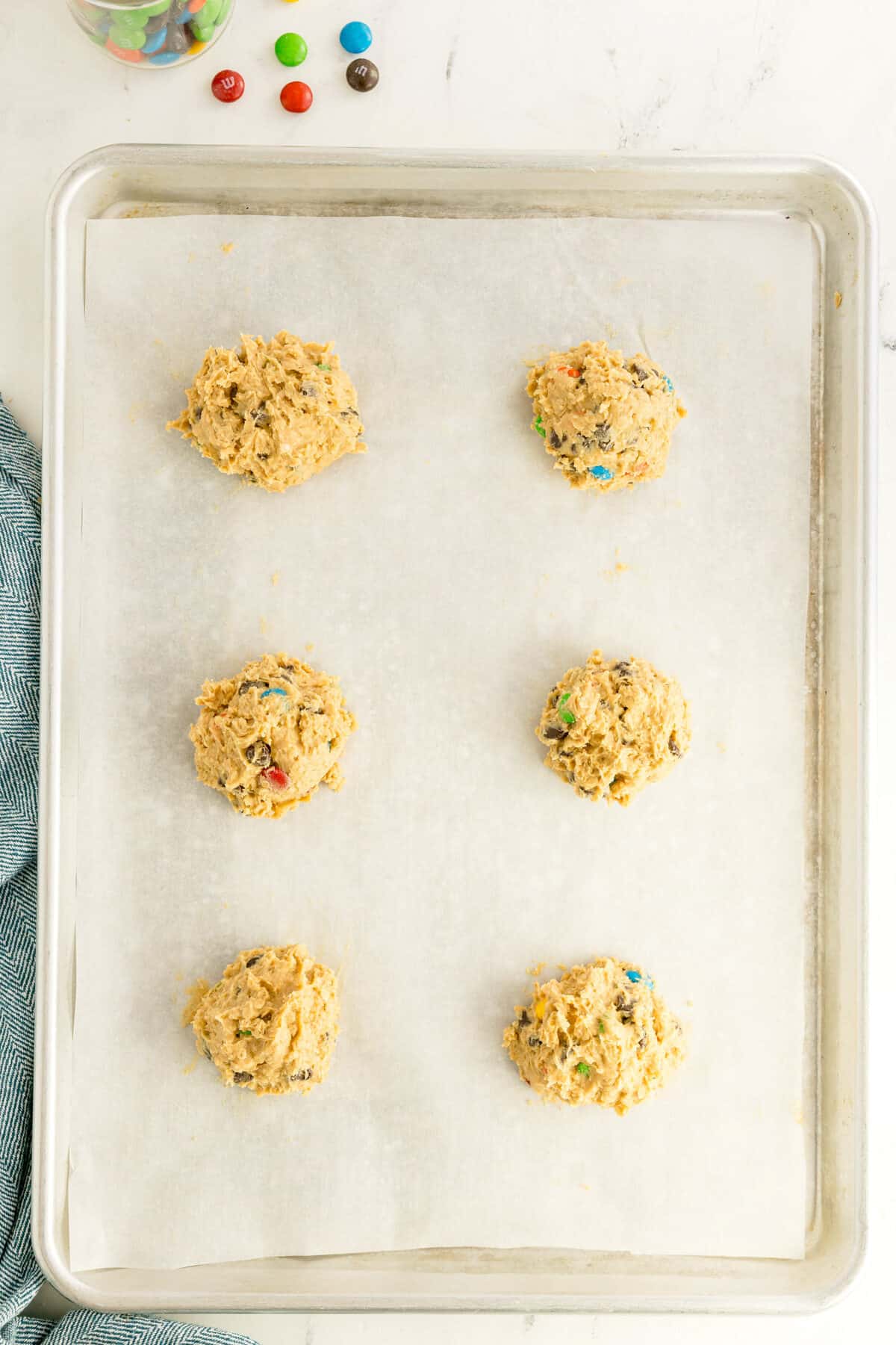 A baking sheet lined with parchment paper holds six scoops of cookie dough with colorful candy pieces, ready to be baked. A blue cloth and a glass jar with candies are nearby.