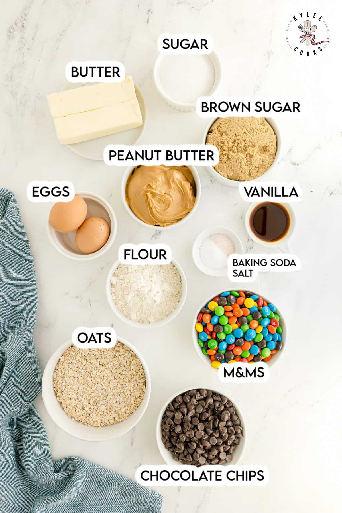 Top-down view of labeled baking ingredients in bowls, including butter, sugar, brown sugar, eggs, peanut butter, flour, vanilla, oats, baking soda, M&Ms, and chocolate chips.