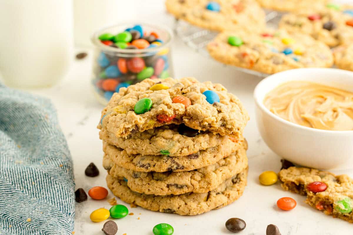 A stack of oatmeal cookies with colorful candy pieces sits on a white surface, surrounded by a bowl of peanut butter, a jar of candies, chocolate chips, and a glass of milk.