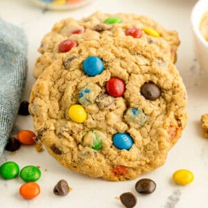 Close-up of two oatmeal cookies with colorful candy-coated chocolates and chocolate chips, resting on a light surface with candy pieces scattered nearby.