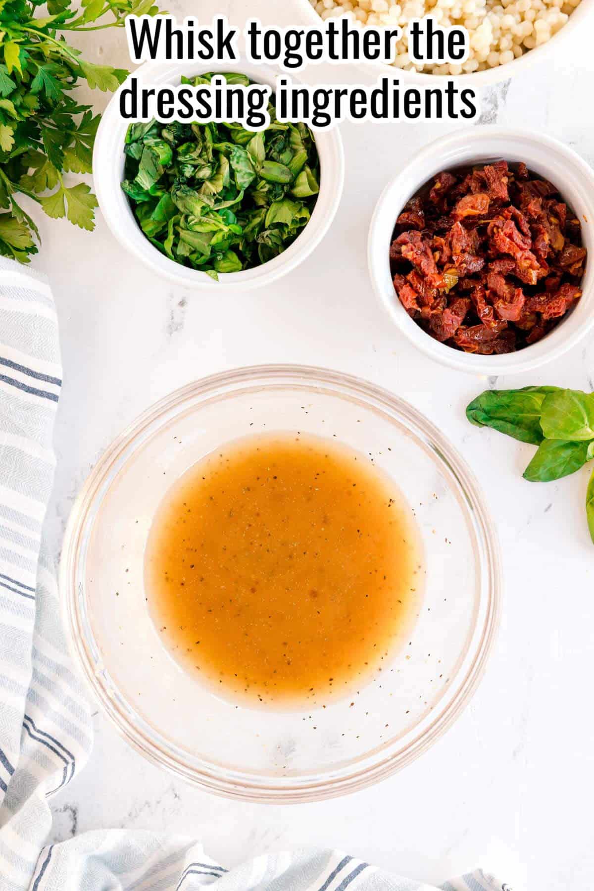 A glass bowl with whisked salad dressing sits on a white surface near bowls of chopped greens and sun-dried tomatoes, ready for a refreshing Summer Couscous Salad. Text above reads, "Whisk together the dressing ingredients.