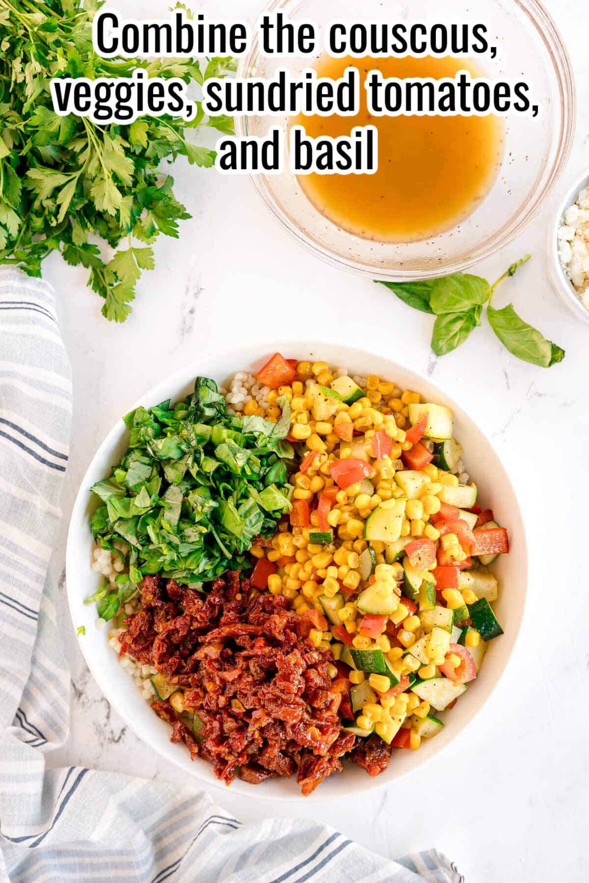 A bowl with Summer Couscous Salad, featuring couscous, chopped vegetables, sundried tomatoes, and basil, sits beside a plate of dressing, fresh herbs, and a striped cloth.