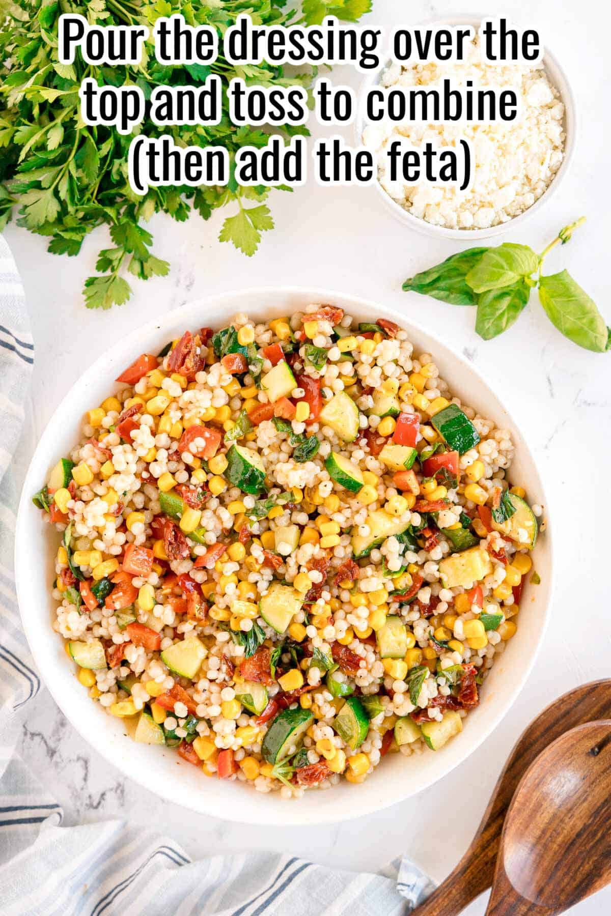 A bowl of Summer Couscous Salad with chopped vegetables sits on a marble surface, surrounded by parsley, basil, a bowl of feta, and wooden utensils. Text reads, "Pour the dressing over the top and toss to combine (then add the feta).