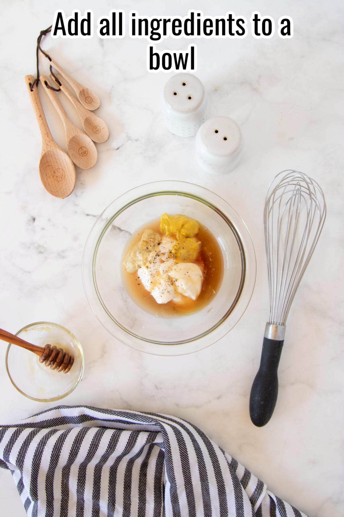 A glass bowl filled with homemade honey mustard dipping sauce sits on a marble counter, surrounded by measuring spoons, salt and pepper shakers, a whisk, a striped towel, and a honey jar.