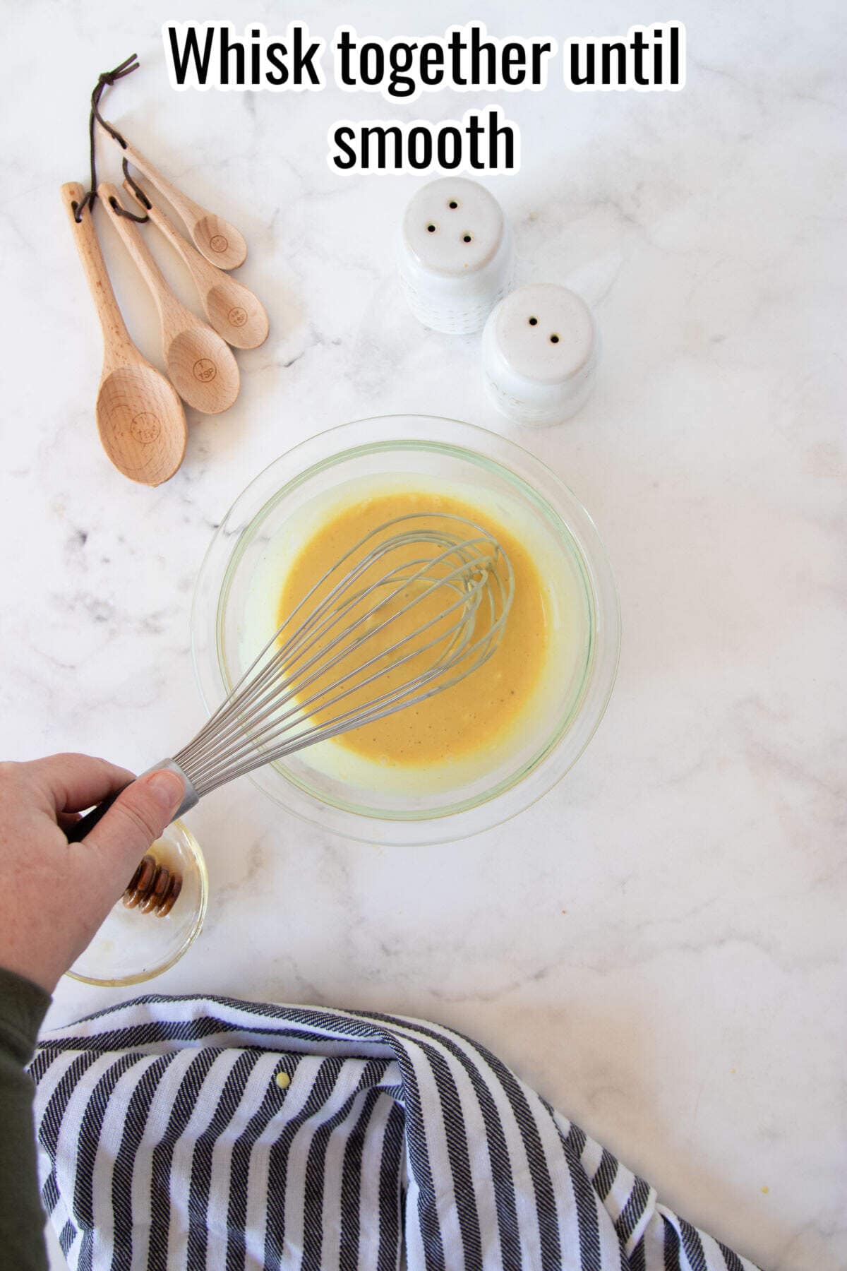 A hand uses a whisk to mix yellow honey mustard batter in a glass bowl on a white countertop, with wooden spoons, salt and pepper shakers, and a striped towel nearby. Text reads "Whisk together until smooth for your dipping sauce.