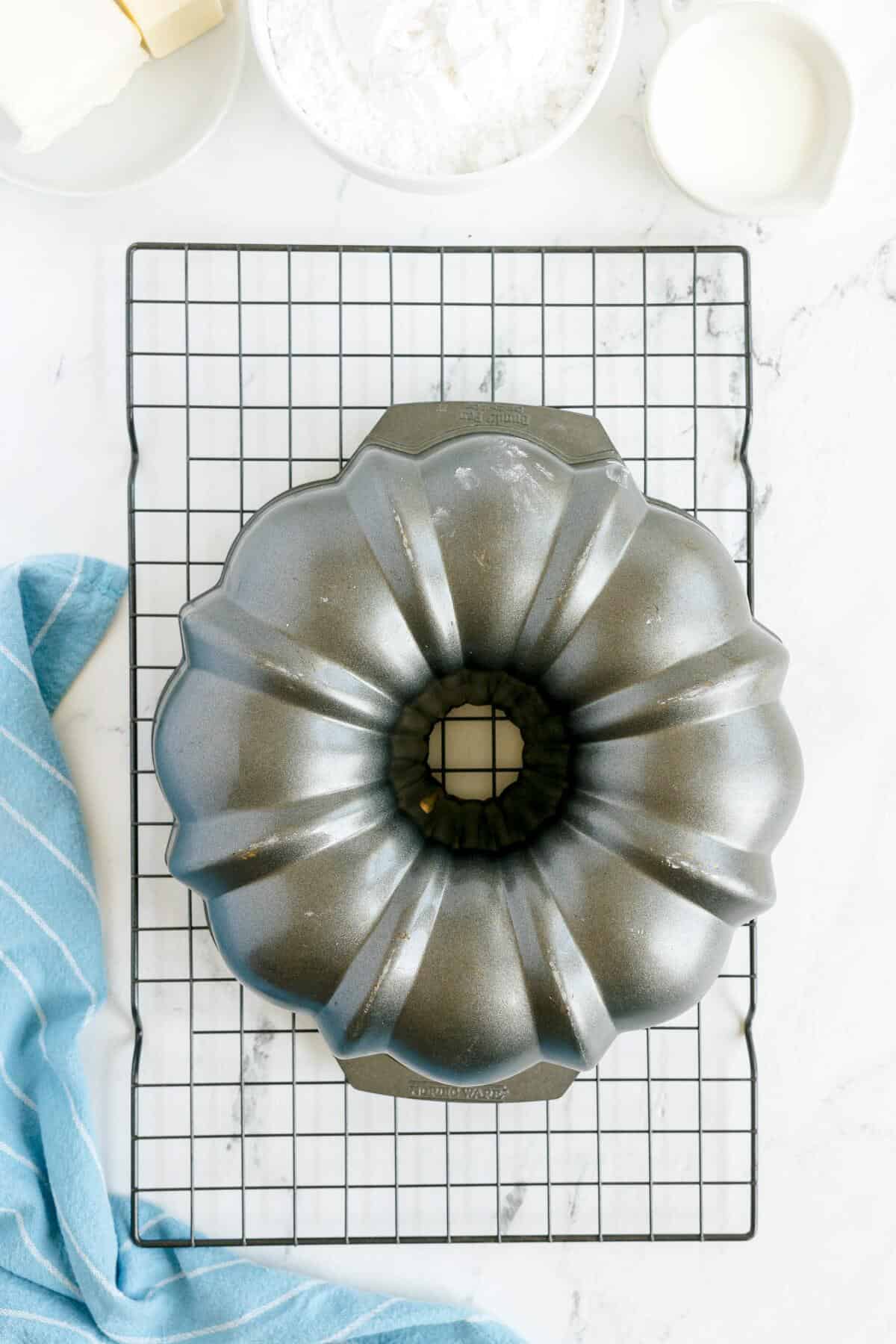 A metal Bundt pan sits upside down on a cooling rack, with a blue striped towel and baking ingredients visible nearby.