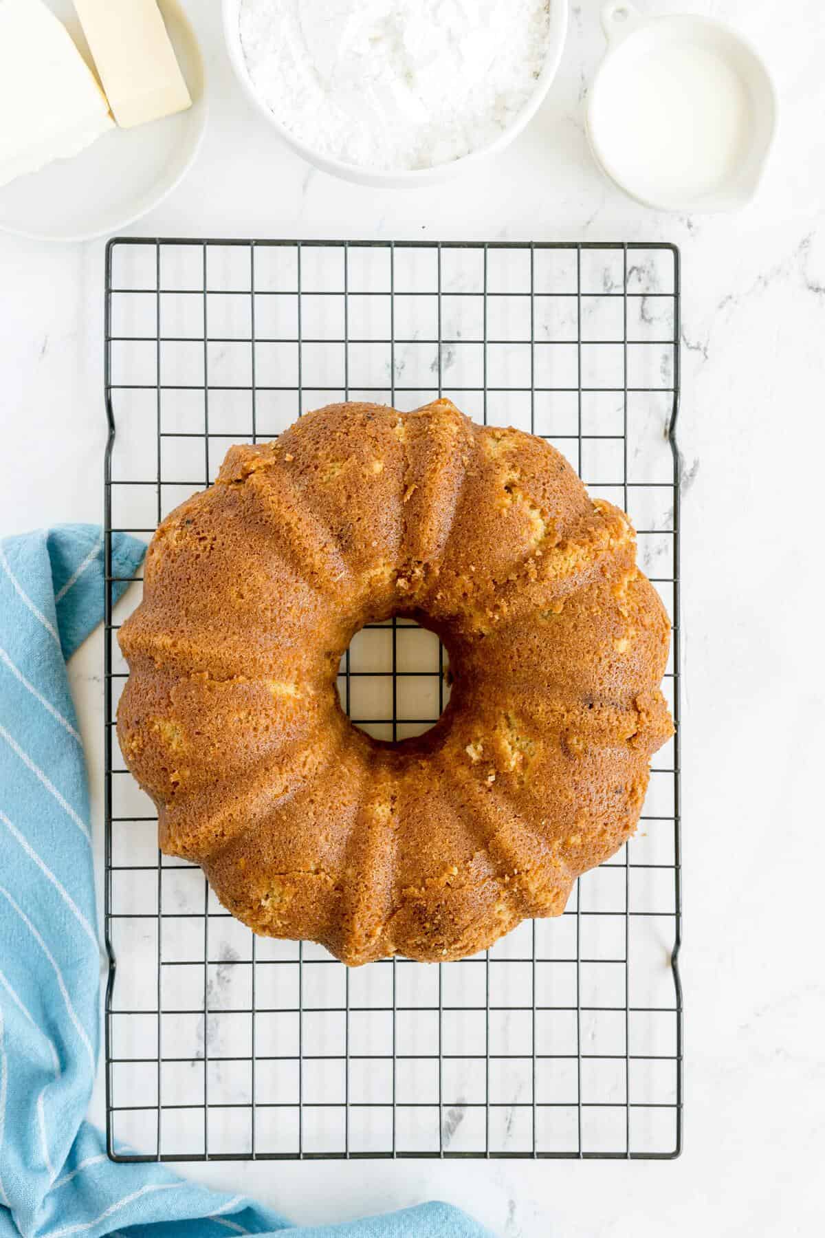 A golden brown bundt cake cooling on a black wire rack with a blue kitchen towel beside it and bowls of ingredients in the background.