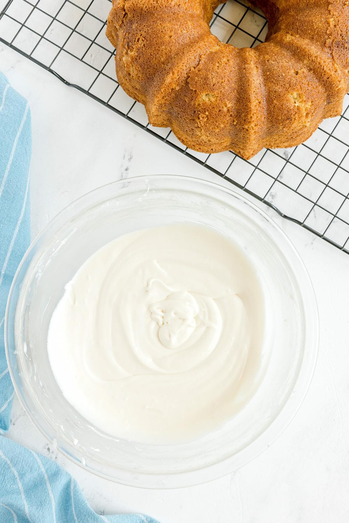 A bowl of white icing sits on a counter next to a Bundt cake cooling on a wire rack.