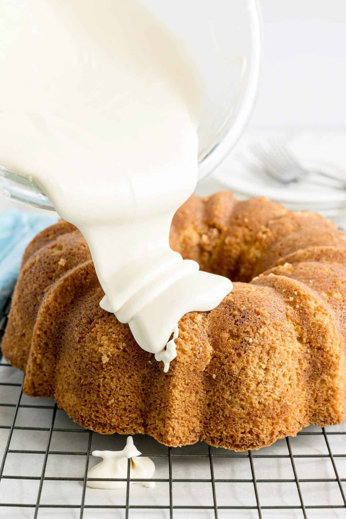 A glass bowl pours white icing over a plain bundt cake on a cooling rack.