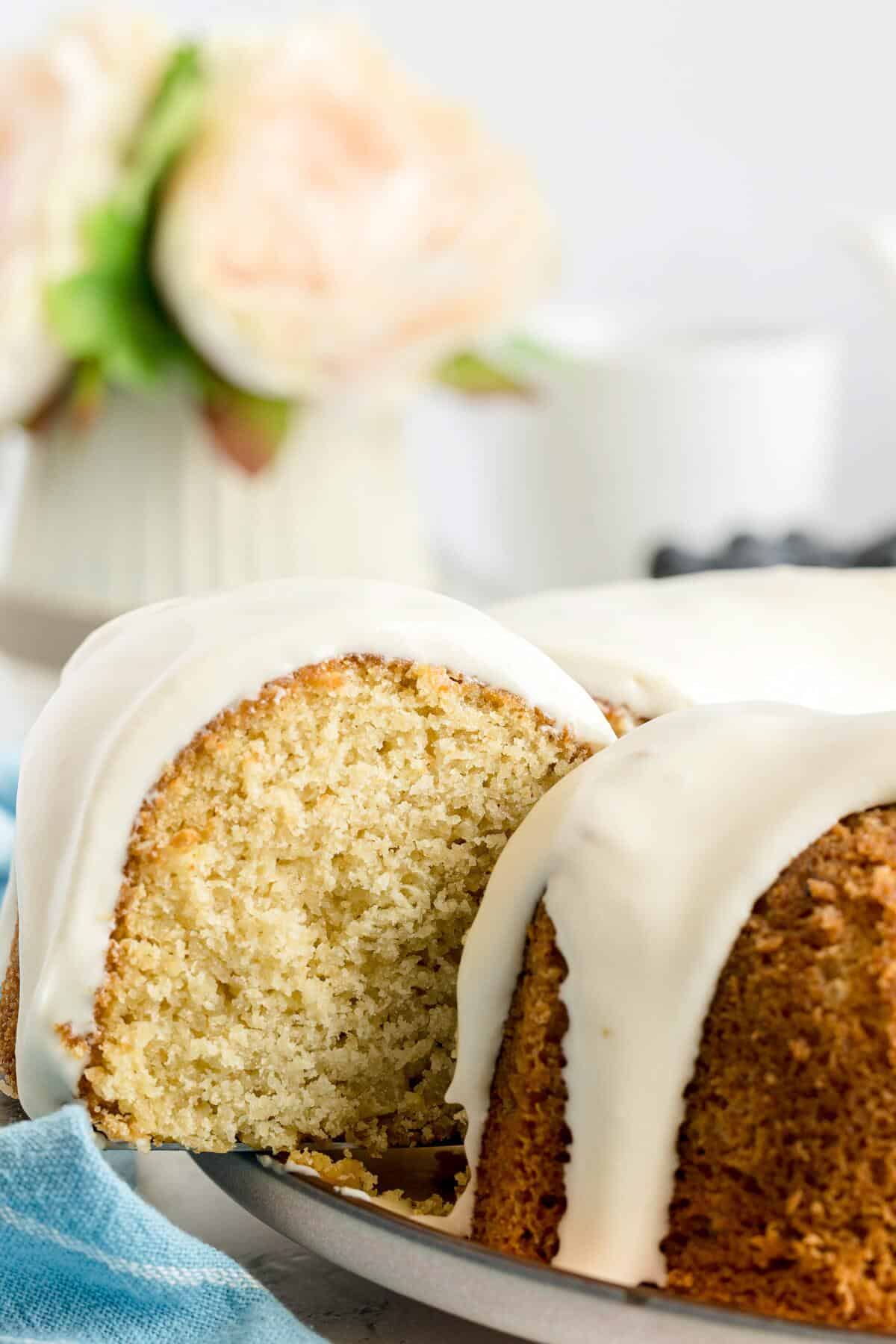 A sliced bundt cake with white icing sits on a plate, with a blurred vase of flowers and a white mug in the background.