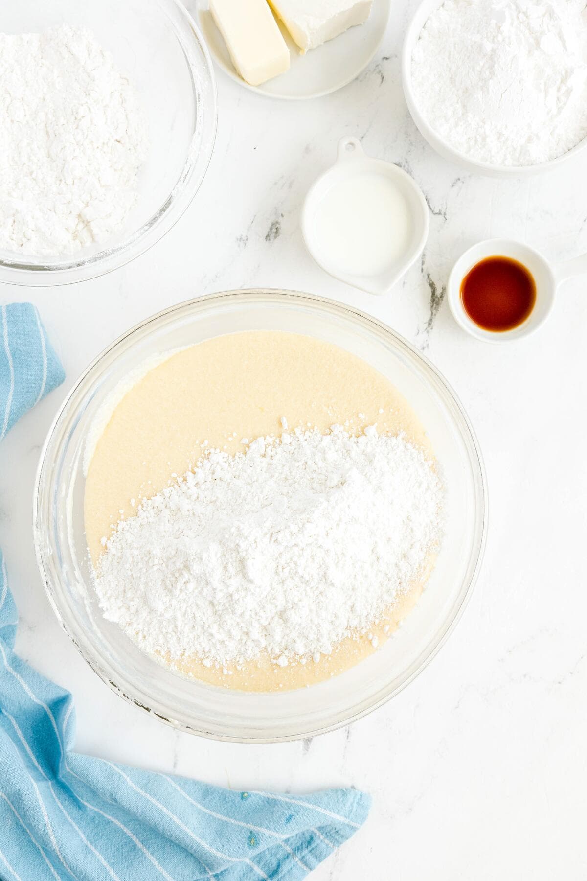 A bowl with creamed ingredients is topped with flour, surrounded by more flour, butter, milk, and vanilla in small bowls on a white countertop with a blue cloth.