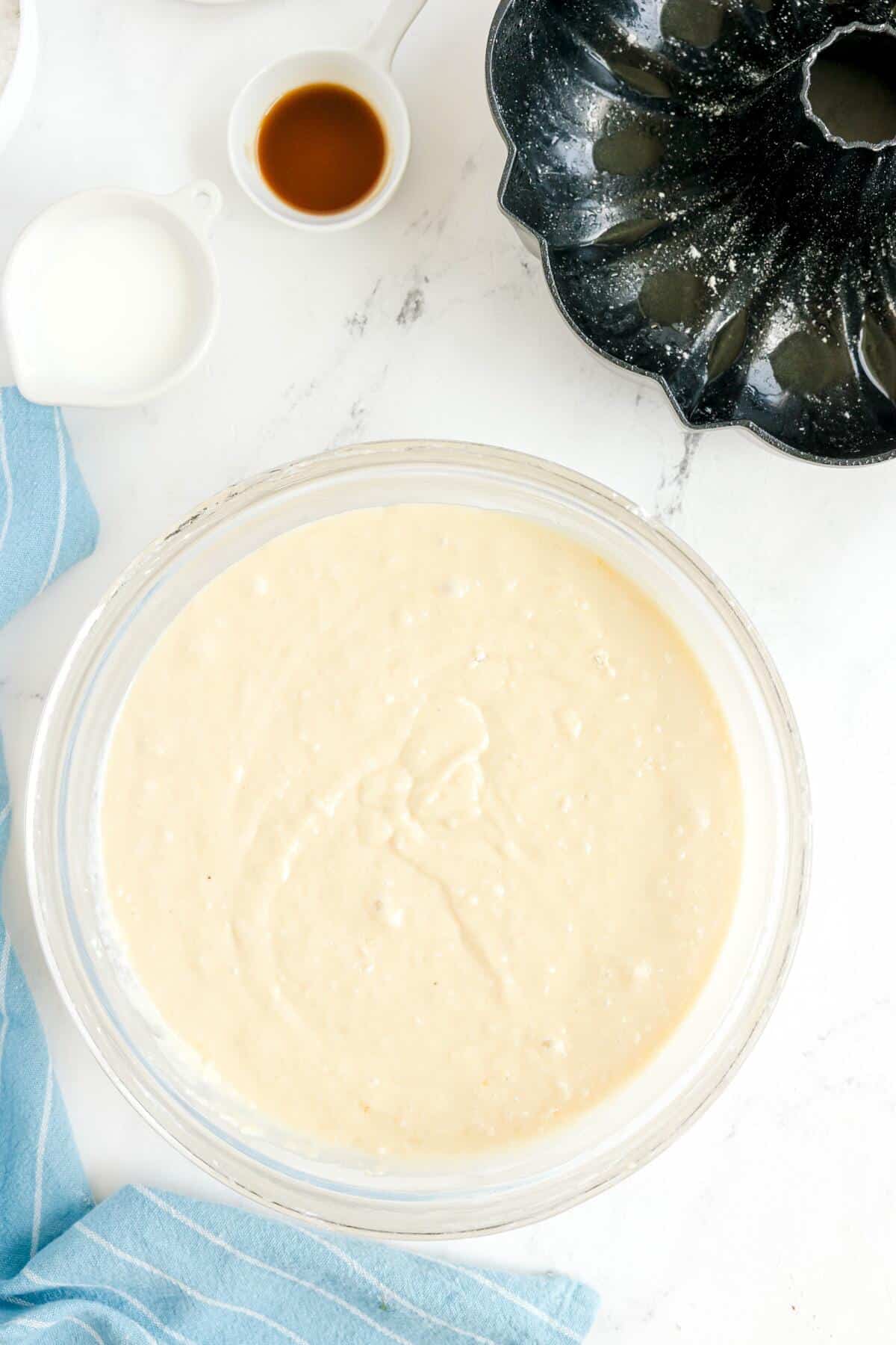 Large glass bowl filled with cake batter on a white surface, surrounded by a bundt pan, small cups of milk and vanilla extract, and a blue striped towel.