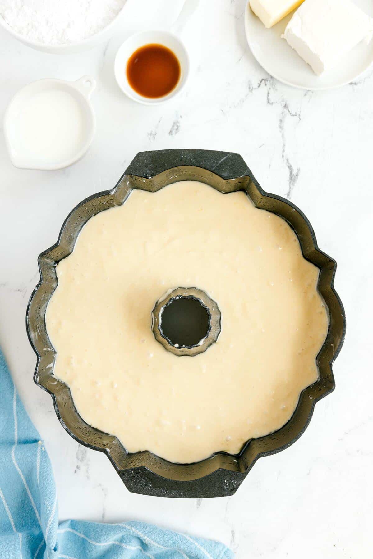 A bundt pan filled with cake batter on a marble surface, surrounded by a bowl of flour, a cup of milk, a plate with butter and cream cheese, vanilla extract, and a blue towel.
