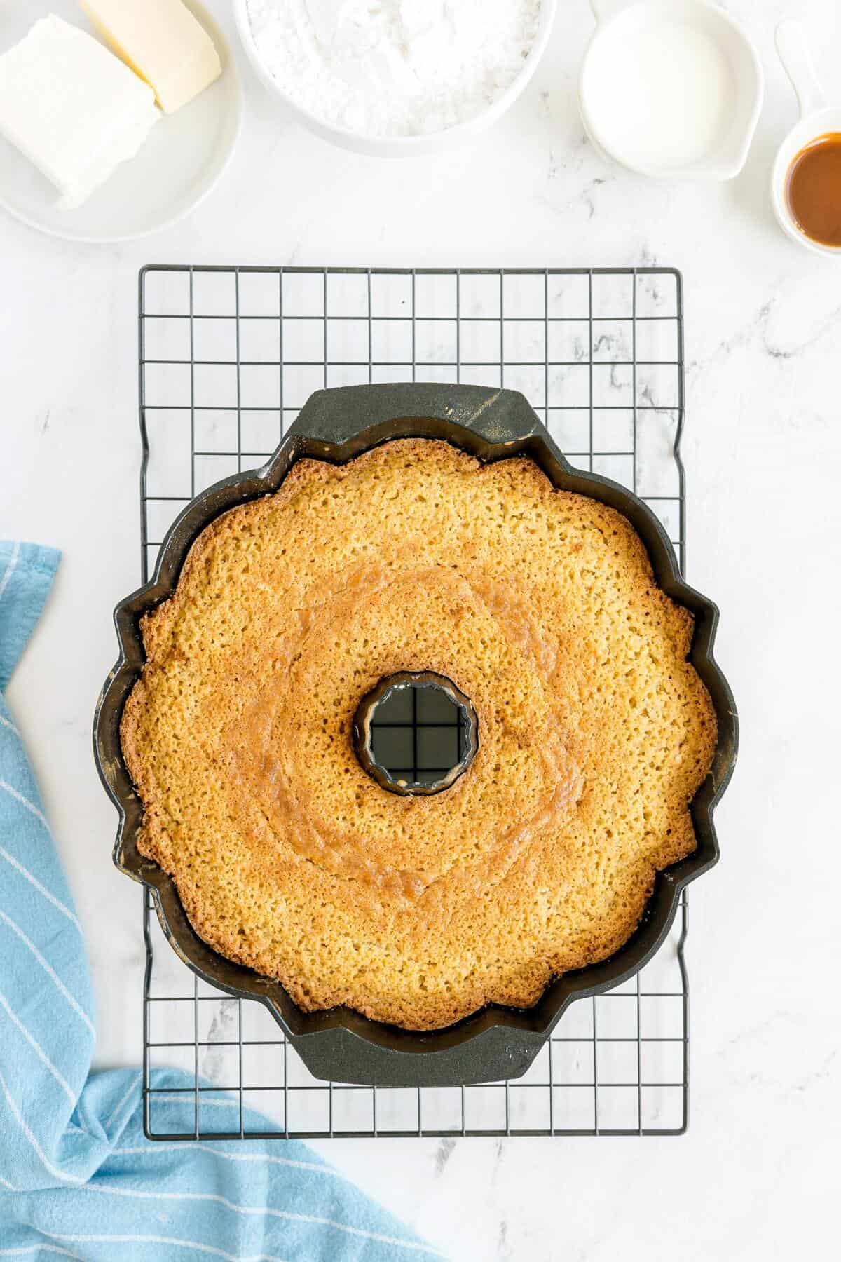 A freshly baked bundt cake cools in a scalloped-edge pan on a wire rack, with baking ingredients and a blue towel nearby.