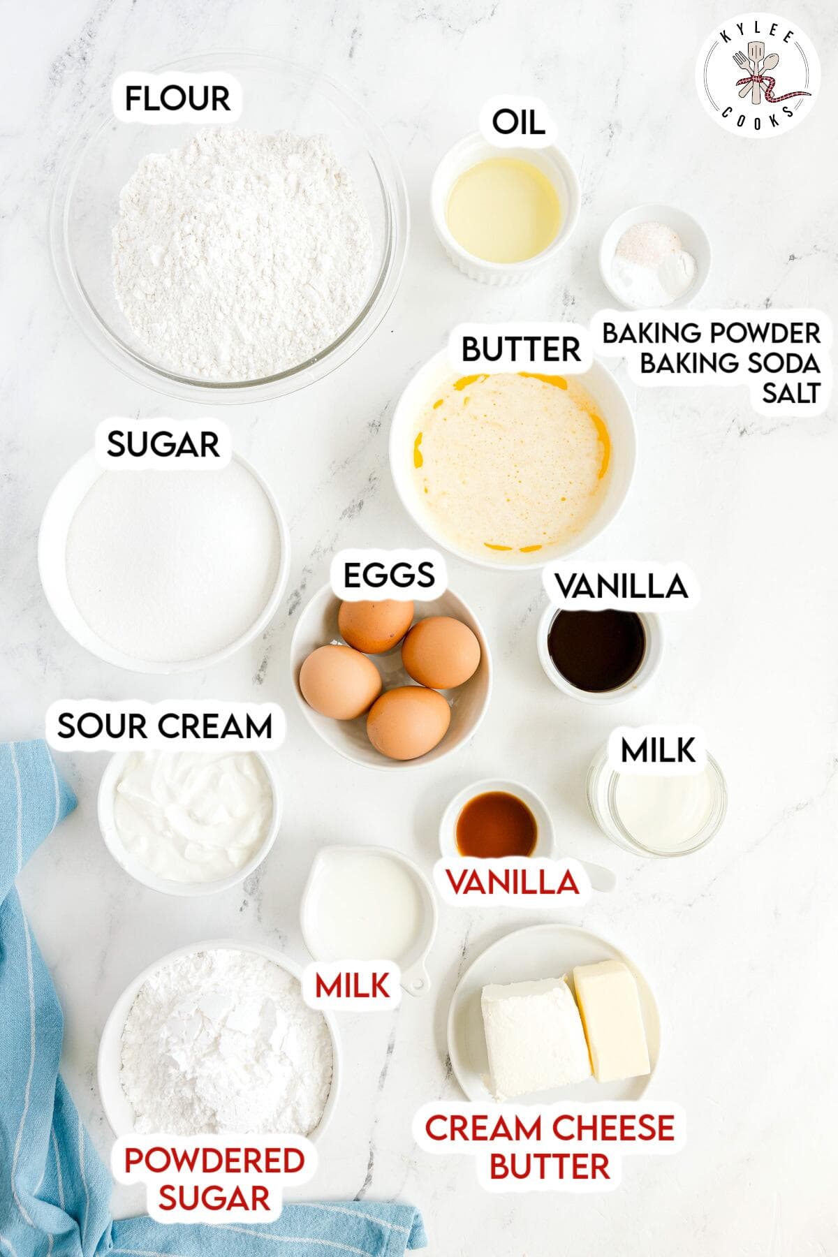 Top-down view of labeled baking ingredients in bowls and containers, including flour, sugar, eggs, sour cream, oil, butter, milk, vanilla, cream cheese, powdered sugar, baking powder, baking soda, and salt.