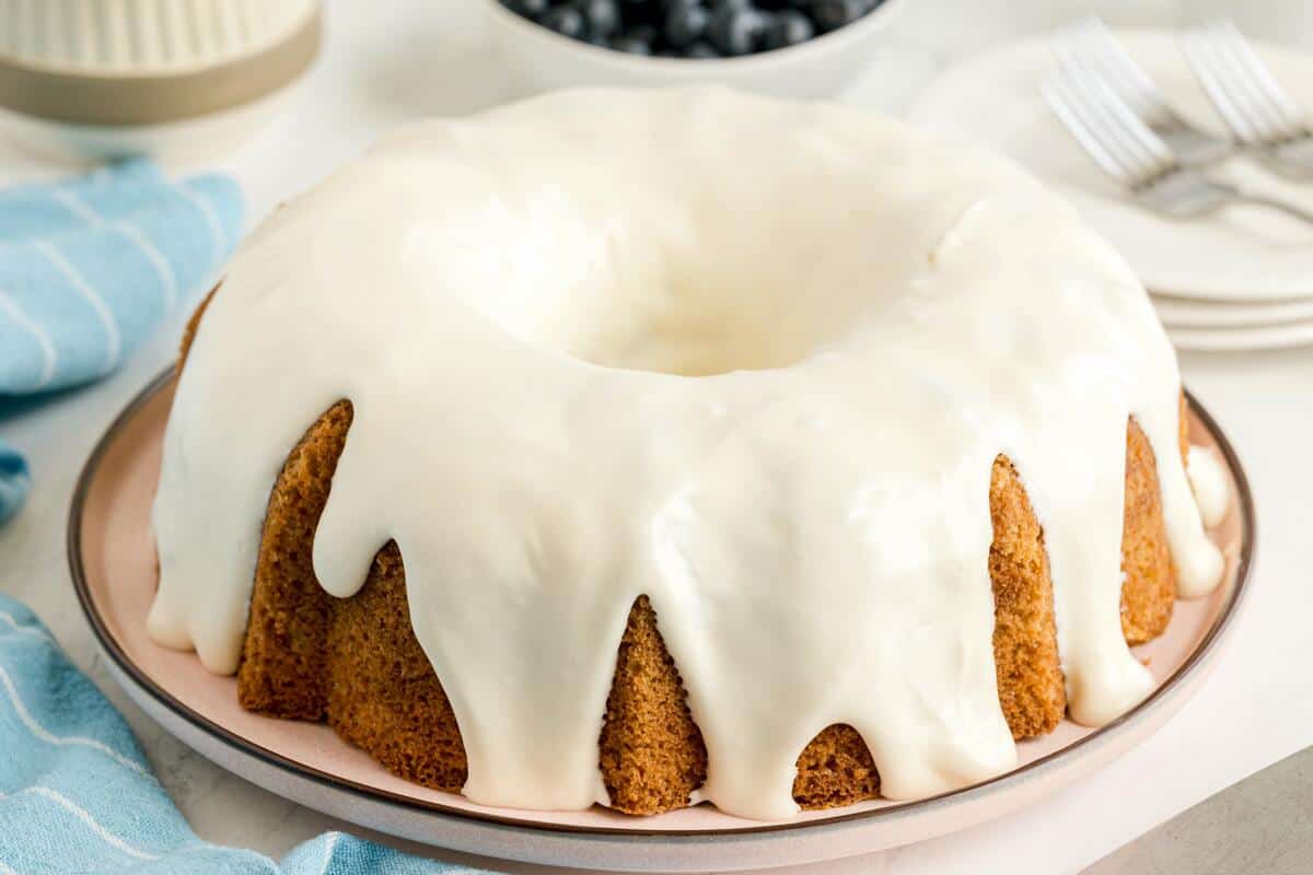A round bundt cake with white icing glaze sits on a plate, with a bowl of blueberries and forks in the background.